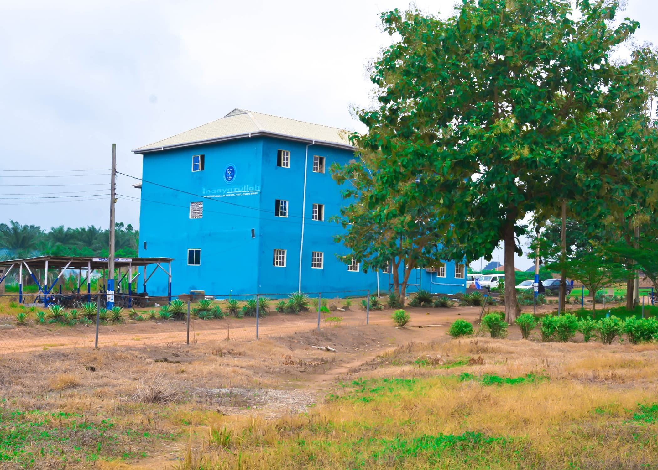 A blue Inaayatullah Academy building surrounded by trees and landscaped grounds
