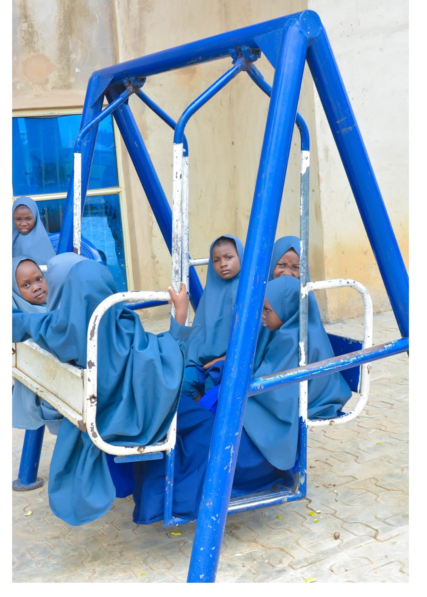 Girls in blue hijabs sitting together on a large blue playground swing
